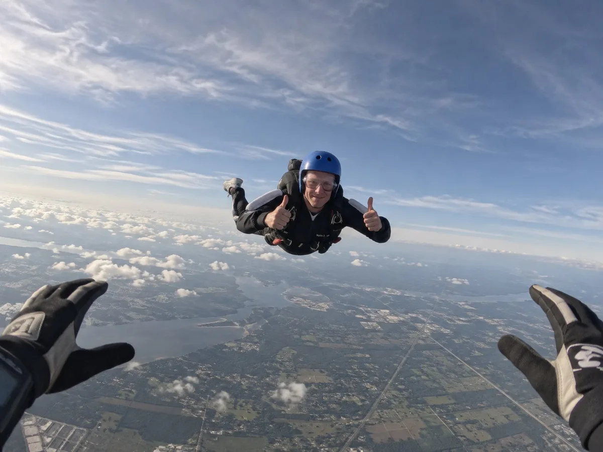 A skydiver in freefall, arms out and grinning at the camera, with fields and cloud far below