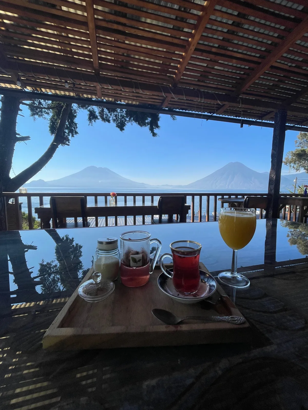 Breakfast on a wooden table under a thatched terrace, with Lake Atitlán and two volcanoes in the background
