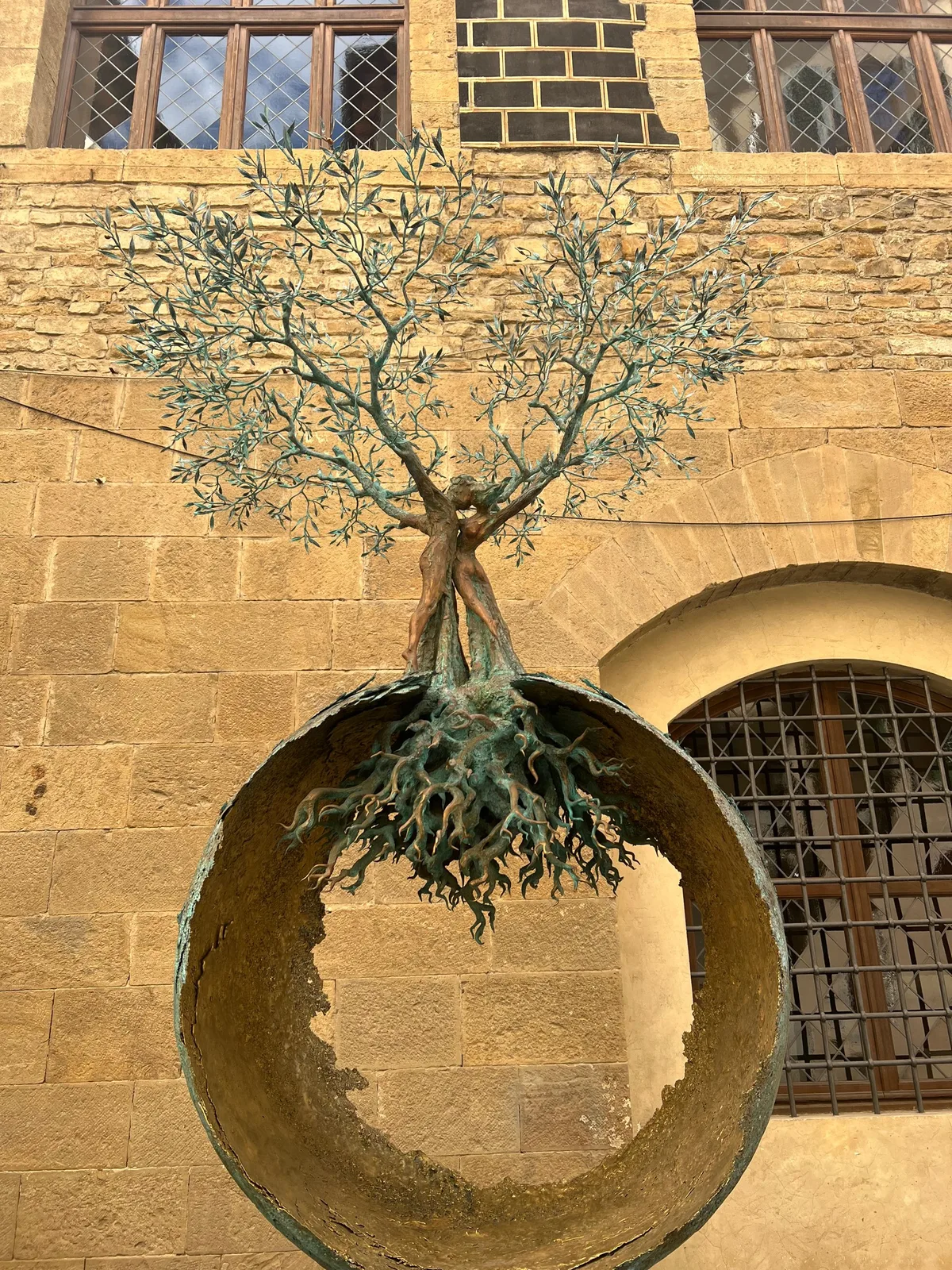A bronze tree with its roots exposed, set into a round stone opening in a wall, against a blue sky