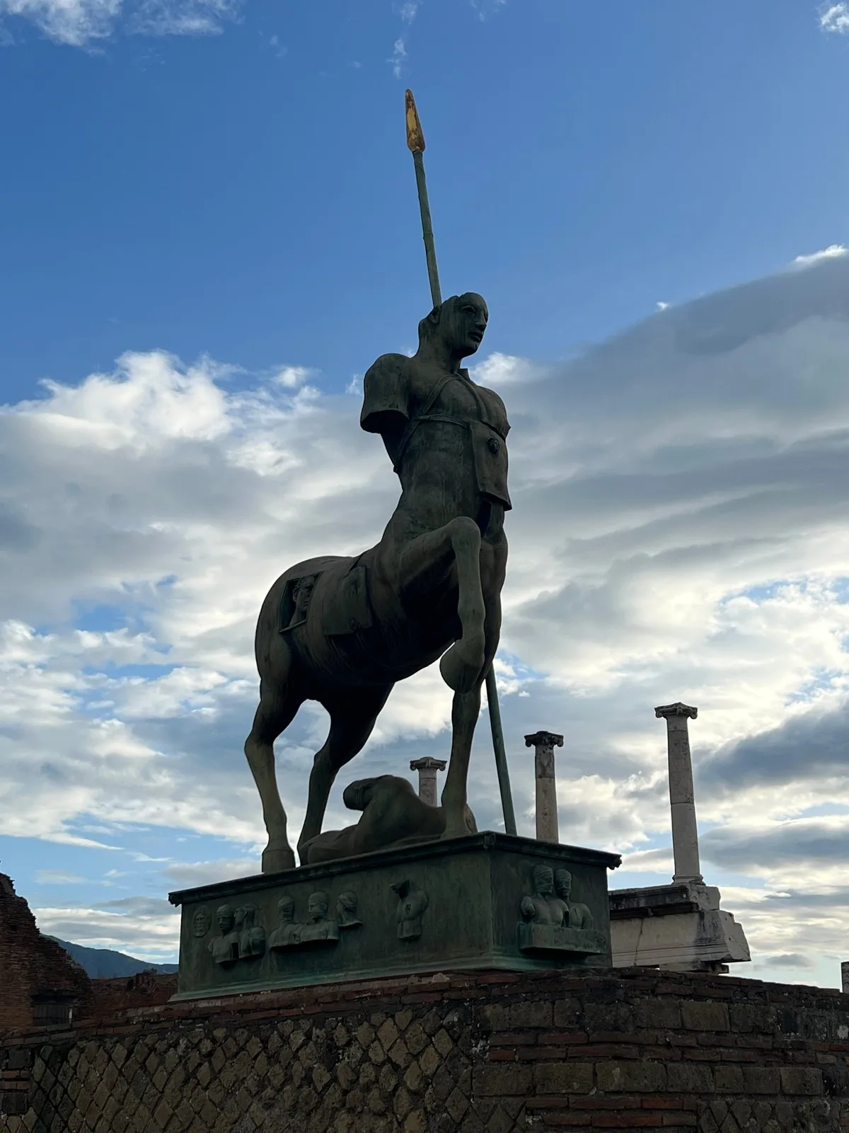 A bronze figure on a tall pedestal silhouetted against dramatic afternoon clouds at Pompeii