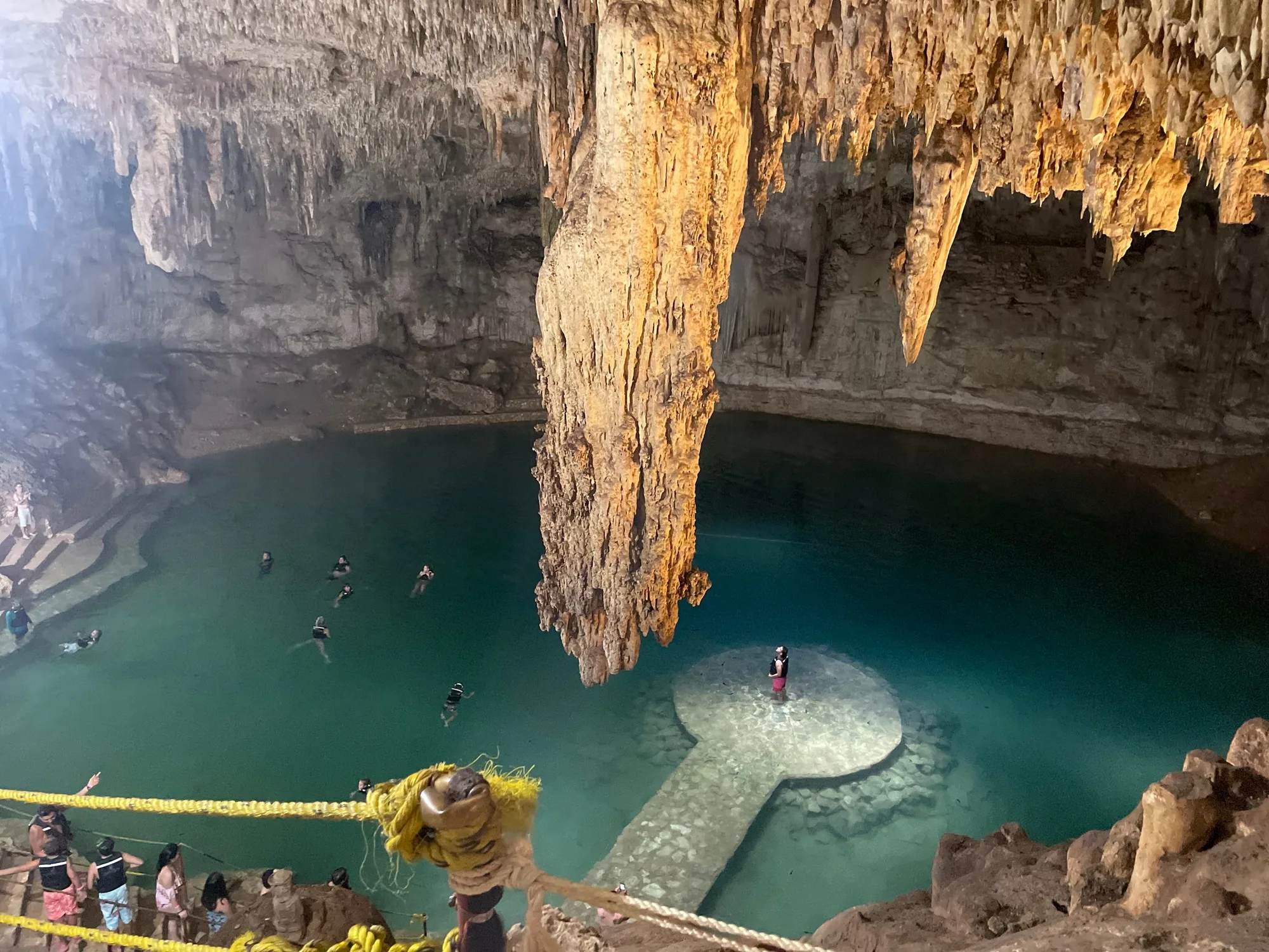 A cenote seen from the rim: a wooden platform over clear turquoise water at the bottom of a limestone cavern, with swimmers below