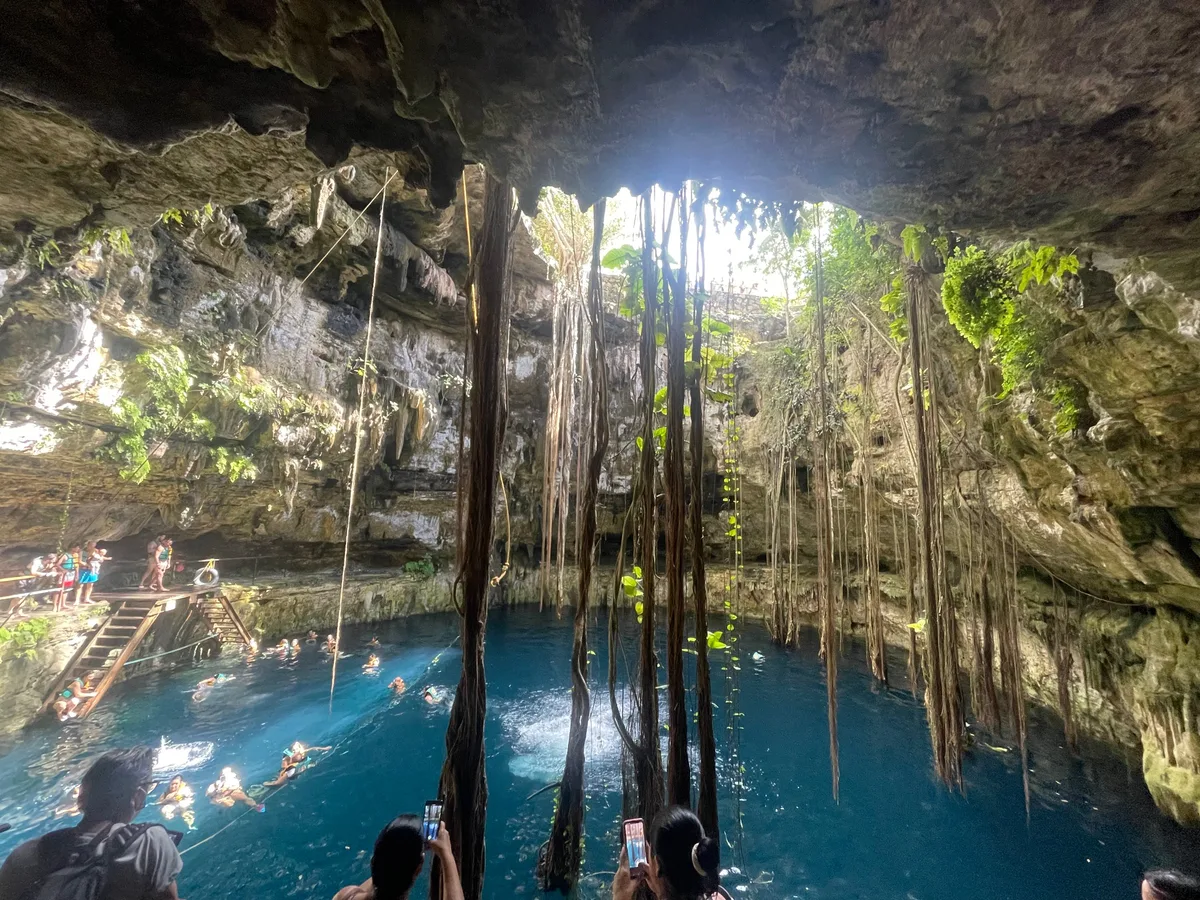 Swimmers in the turquoise water of a cenote, lit by a shaft of daylight from an opening in the cavern roof above