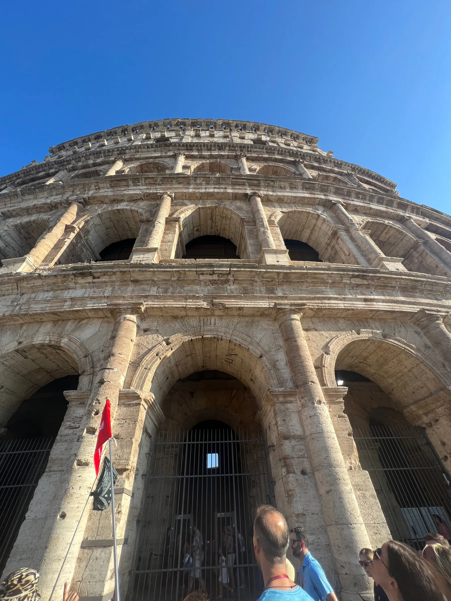 The outer wall of the Colosseum, three tiers of arches, seen from below against a blue sky
