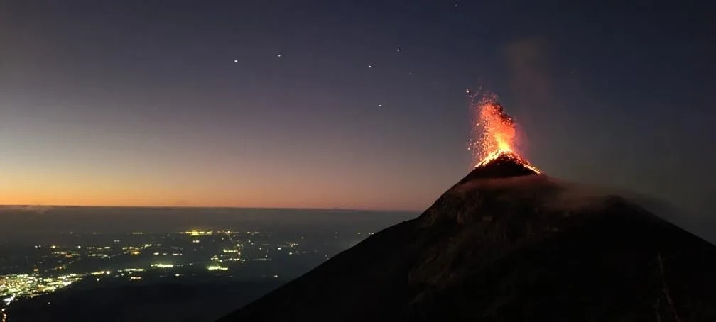 Volcán de Fuego erupting at night, a plume of lava lit orange against a dark blue sky, with the lights of towns spread across the valley below