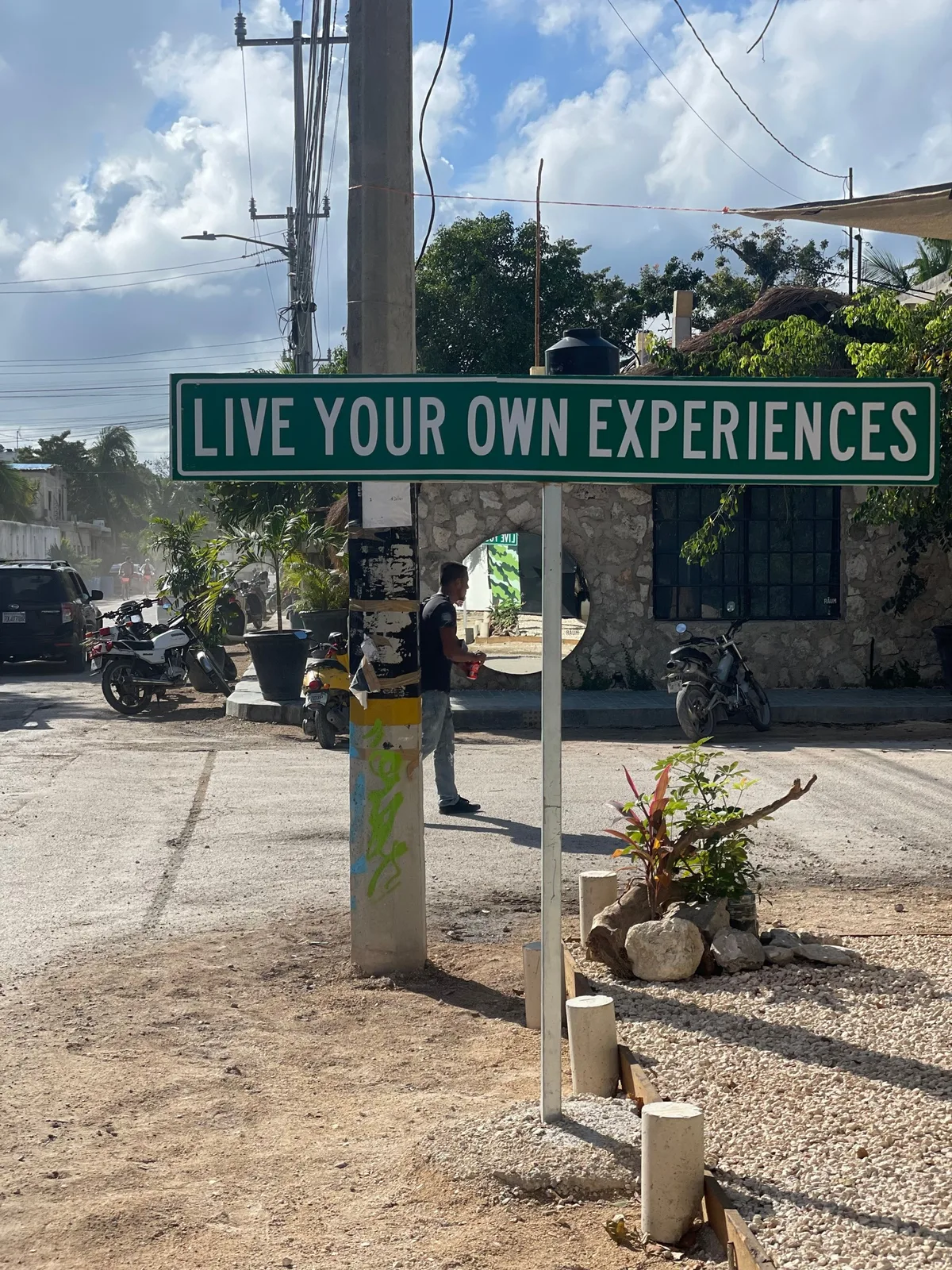 A green street sign reading Live Your Own Experiences on a pole beside a dusty road, with a motorbike and a person nearby