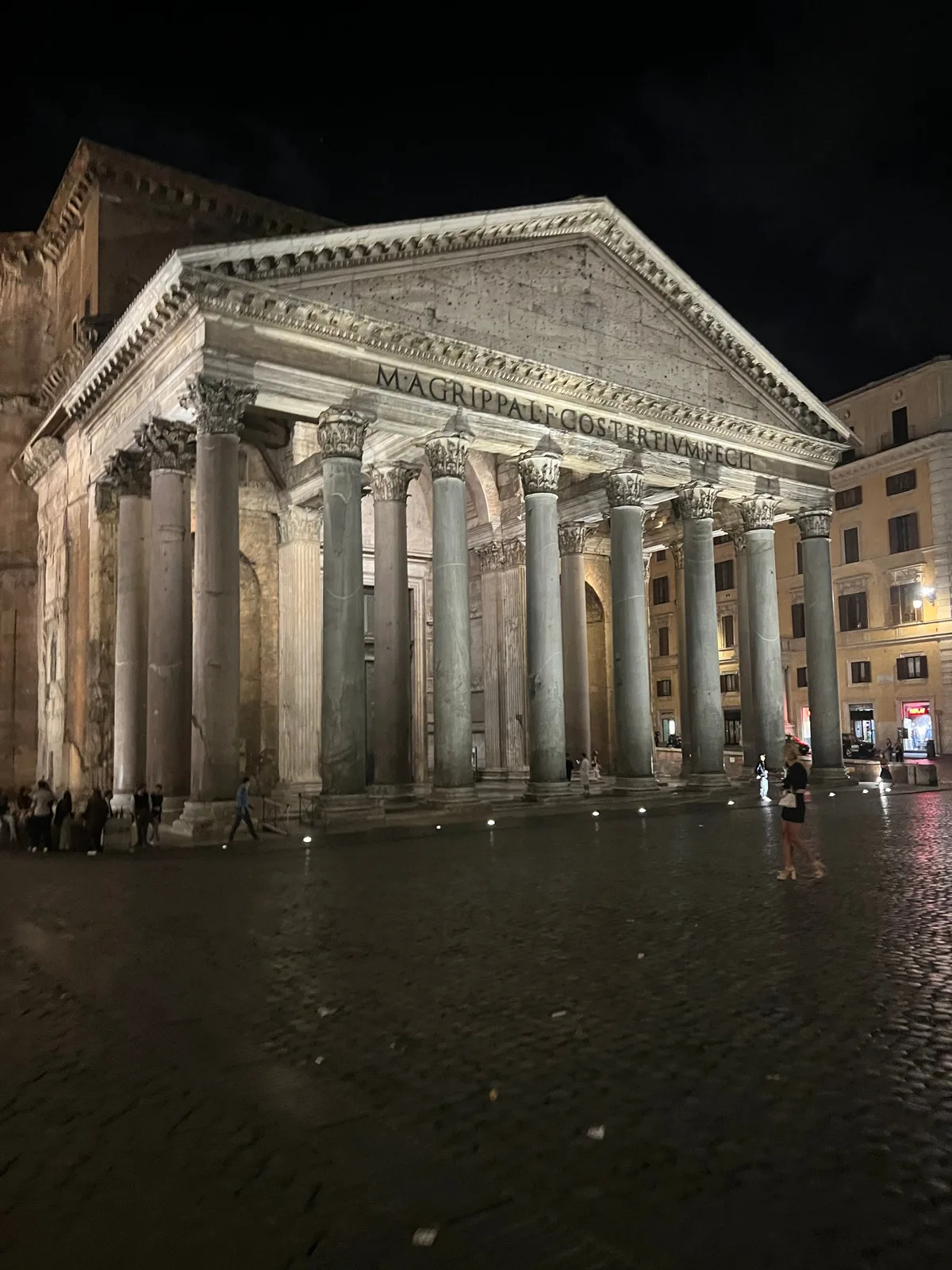 The Pantheon's columned portico lit at night, reflected in the wet stones of an almost empty piazza