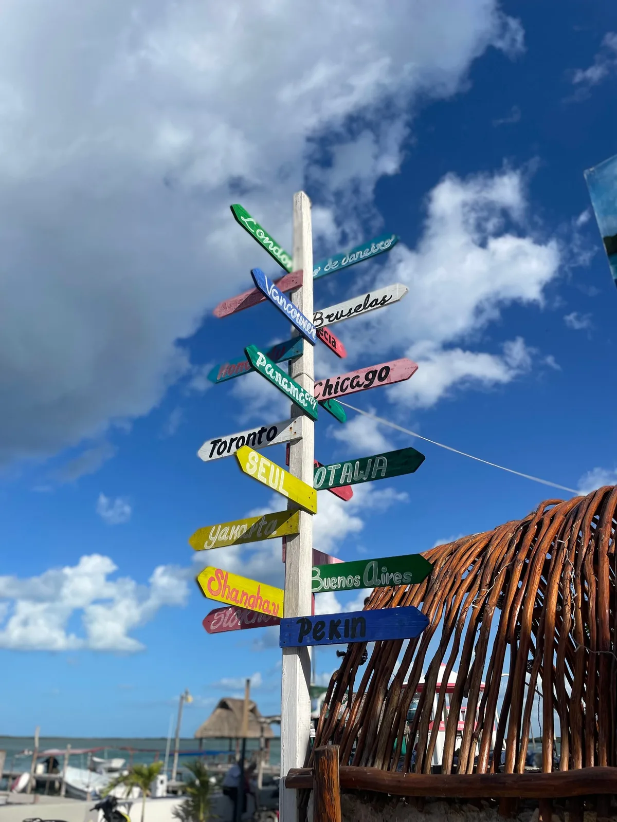 A wooden post covered in colourful arrows pointing to cities around the world, against a blue sky with clouds