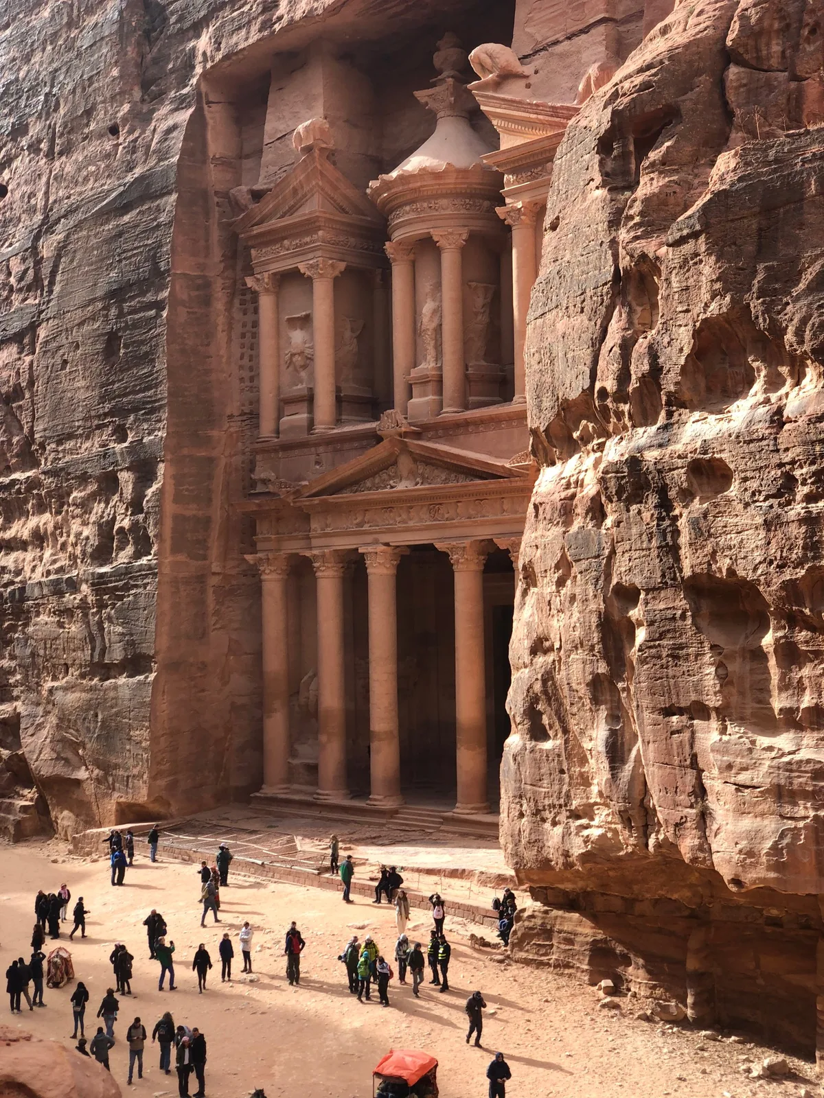 The Treasury at Petra, a columned facade carved into rose-coloured rock, seen from the narrow canyon with visitors small at its base