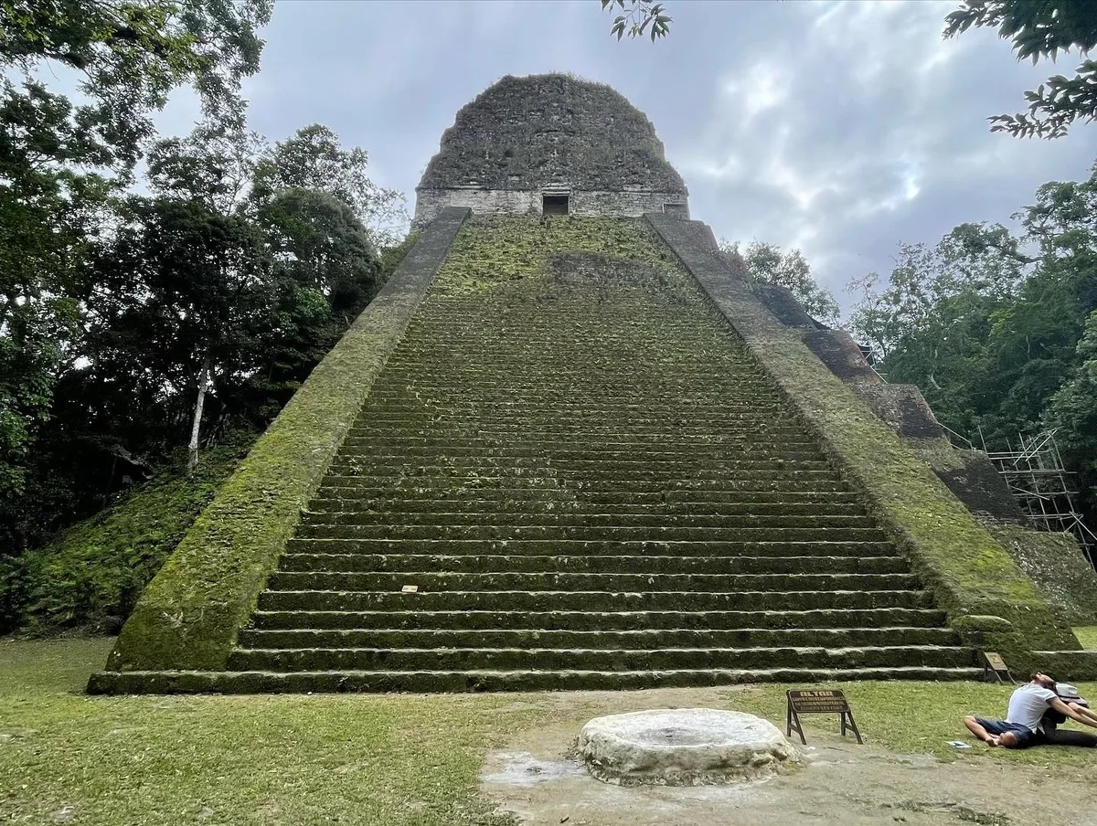 A steep, moss-covered stepped temple at Tikal under a grey sky, with visitors small at its base