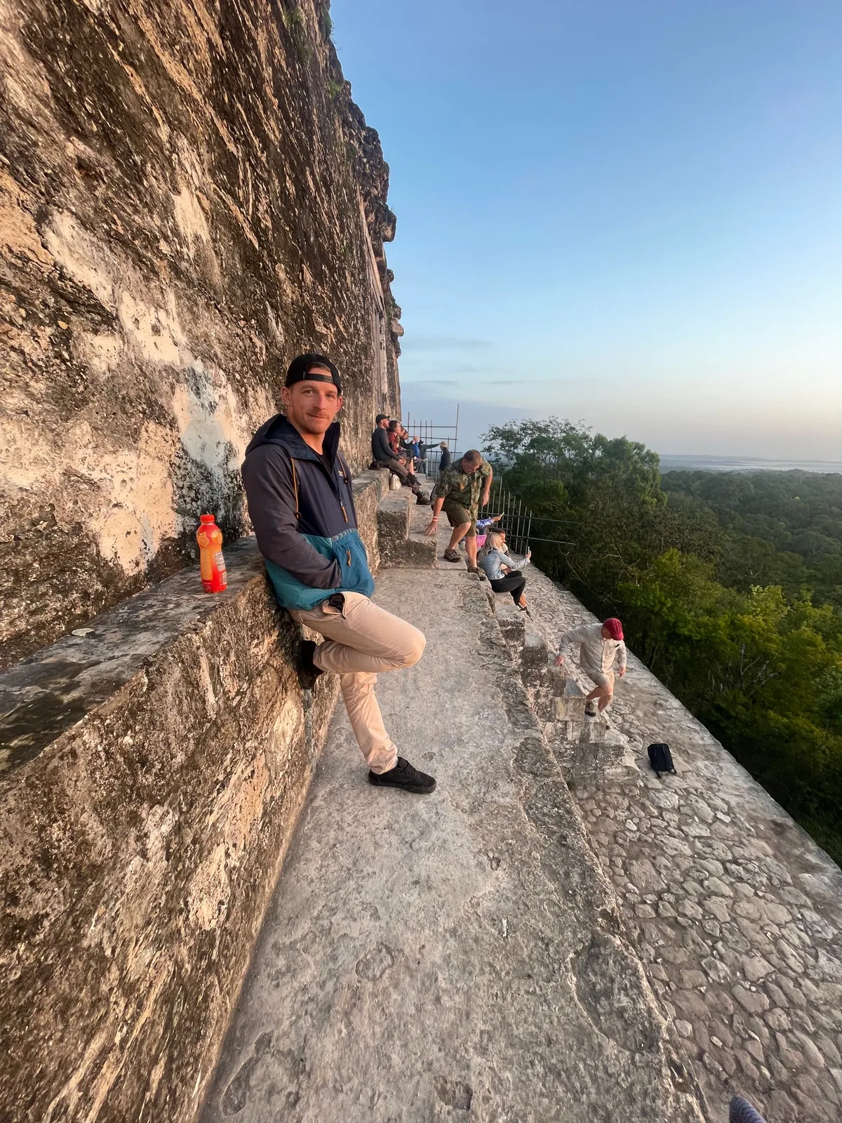 Ryan sitting on the stone edge at the top of a Tikal temple at sunrise, the jungle canopy stretching to the horizon behind him