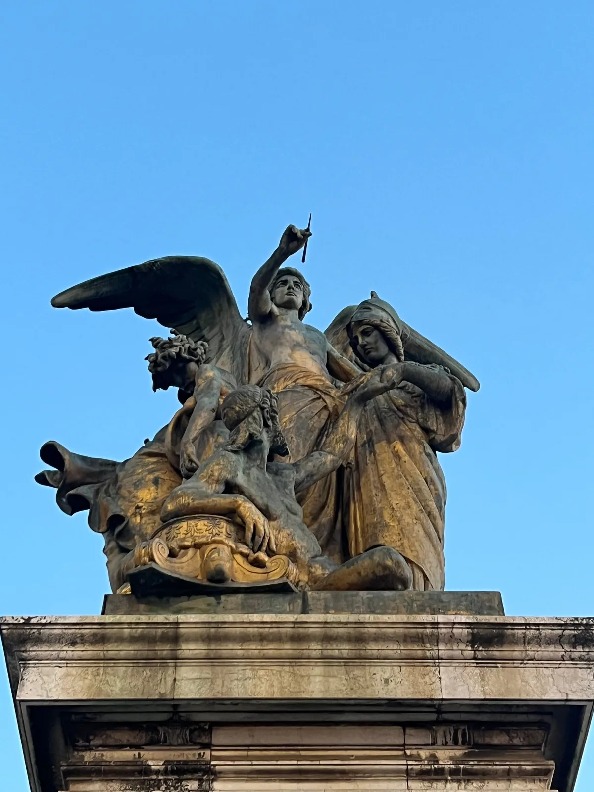 A winged marble figure on top of a stone plinth, photographed from below against a clear sky